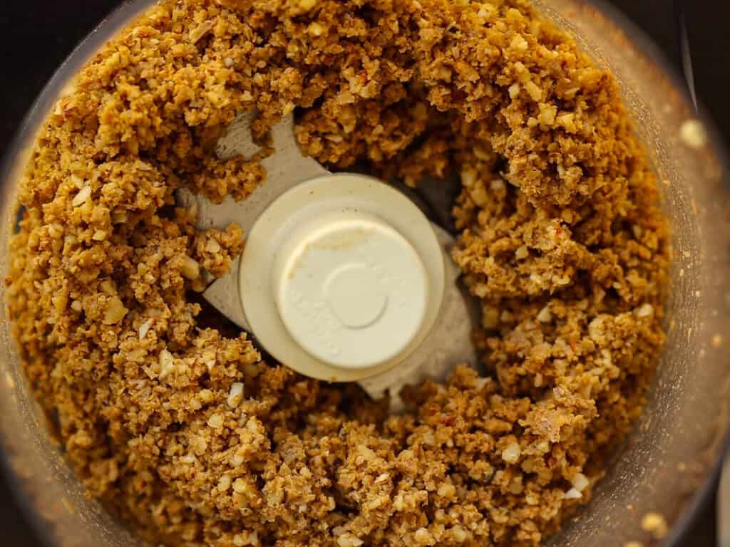 Coarsely ground mixture of nuts and vegan sausage and garlic in a food processor bowl. The blend has a crumbly texture, with visible small bits, and is centered around a white plastic blade.